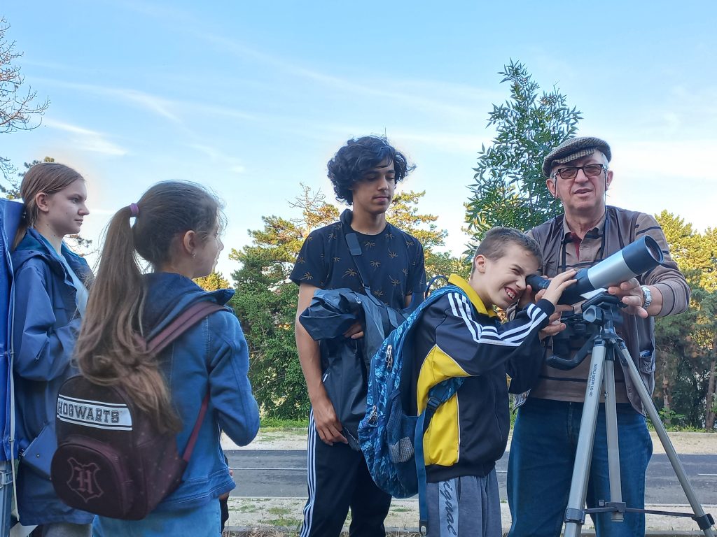 Egy kisfiú éppen belenéz az állványos távcsőbe, a többi diák résztvevő sorban várakozik.