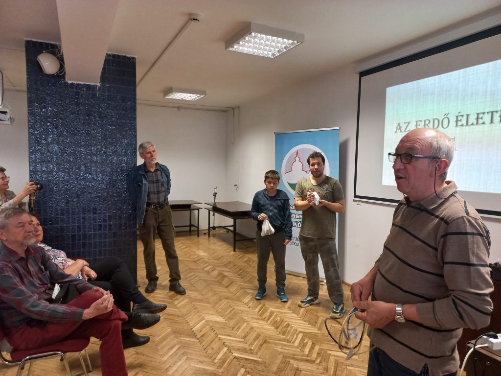 A háttérben a program két résztvevője, egy apuka és egy fiatal fiú tartja a kezében a rigókat tartalmazó vászonzsákokat. A kép előterében dr. Kis Ernő mutatja a közönségnek a kezében tartott, drótra fűzött gyűrűket, amelyekkel a madarakat megjelölik